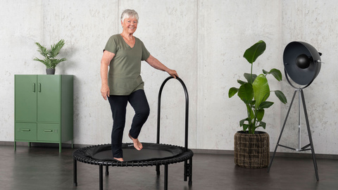Une femme âgée s'entraîne sur le mini-trampoline bellicon et se tient à la barre latérale.