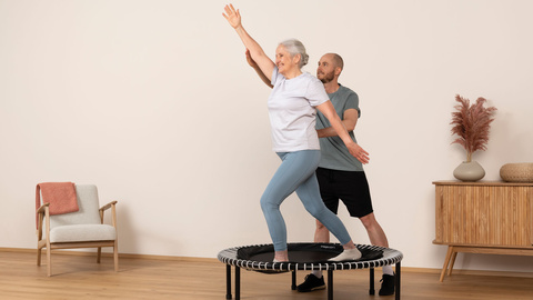 Une dame âgée est accompagnée par un·e physiothérapeute pendant l'entraînement sur le mini-trampoline.